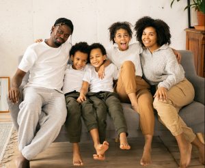 Photo of African American family sitting on couch. From left: father, two sons, daughter and mother on far right. They all have big smiles and have their arms around each other's shoulders. 