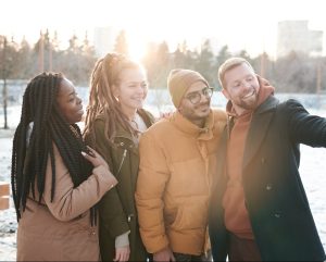 Group Selfie photo. From left: woman with black braided hair, woman with dreads, man with beard and glasses, on far right man with arm stretched out taking the selfie. The group is taking the picture outside in a park during winter. They are all dressed in warm winter coats. 
