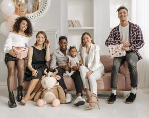 Group photo at a toddler's birthday party. The Group is sitting on a pink couch. From left: woman holding gift, woman seated next to her, father holding female toddler, mother seated to their right, male party guest with gift on far right. 