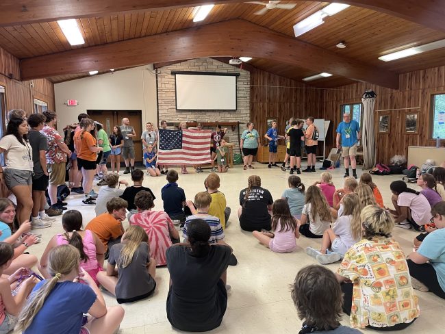 large American flag held by people in an indoor room, with others seated on the floor